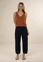 A woman stands against a plain beige studio background wearing a rust-brown ribbed knit sleeveless V-neck top and dark navy wide-leg cropped pants with a drawstring waist, paired with beige flat sandals.